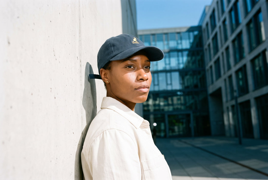 Frau mit marineblauem Kangol Washed Dad Hat lehnt an einer sonnigen Sichtbetonwand in einem Innenhof