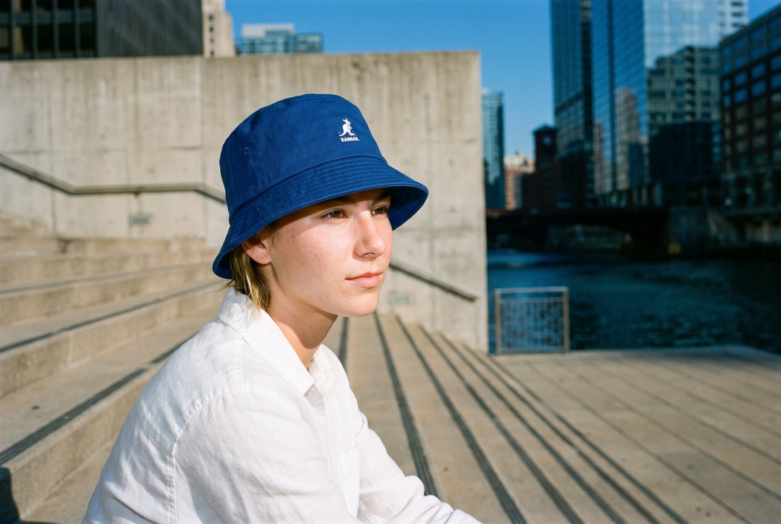 Frau mit royalblauem Kangol Washed Bucket Hat sitzt auf Betonstufen an einem Fluss in der Stadt